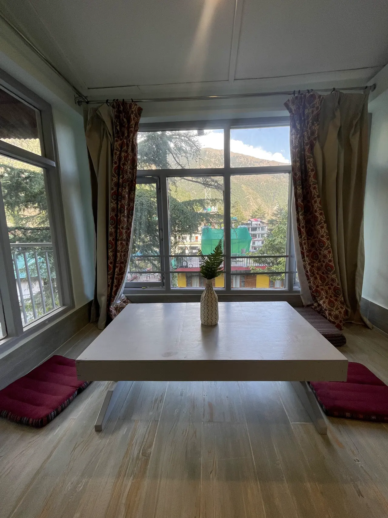 A low table and two floor cushions at the studio's tall windows, trees and a ridge beyond