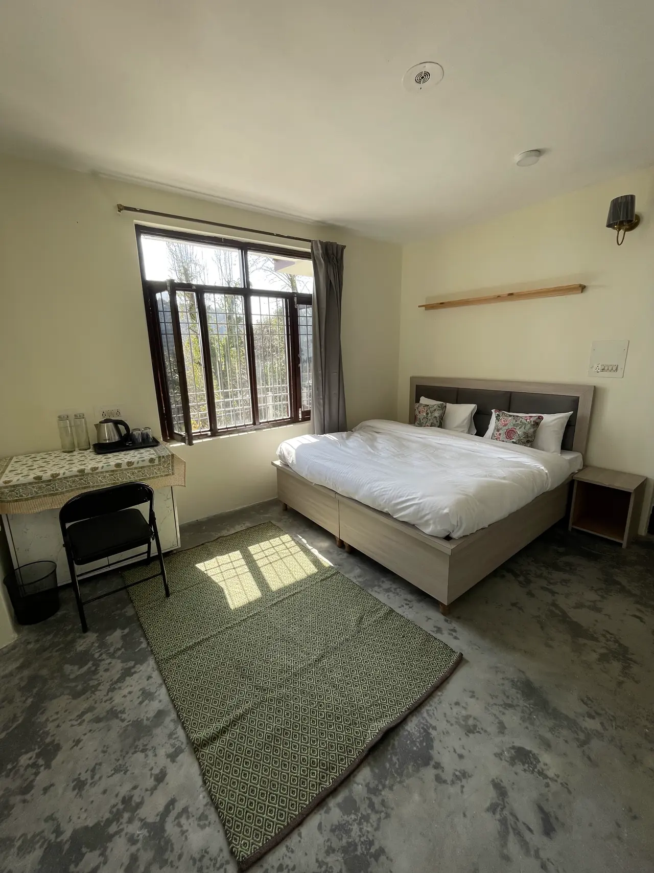 A double bed, wall shelf, green rug and a kettle counter by a window full of trees