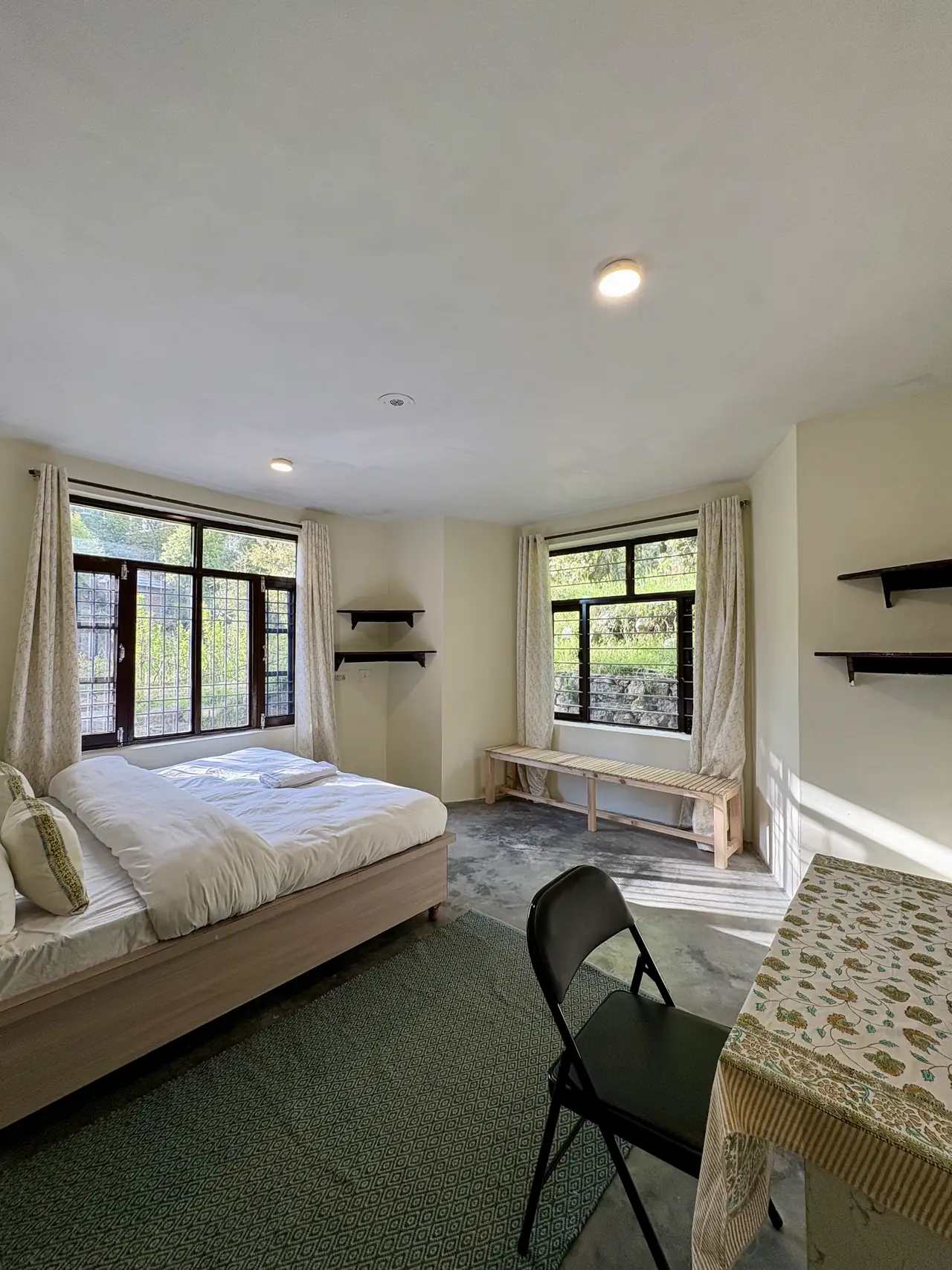A bright room with the bed under one window and a bench under another, greenery outside