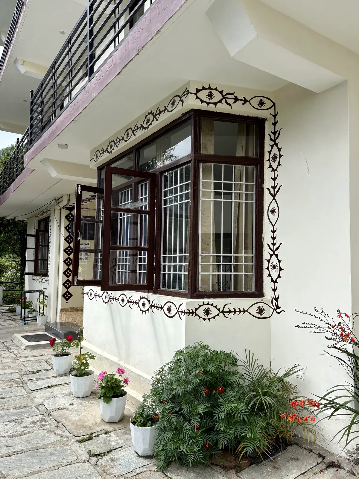 Painted folk motifs framing a bay window on Soma House's white wall, flowering plants below