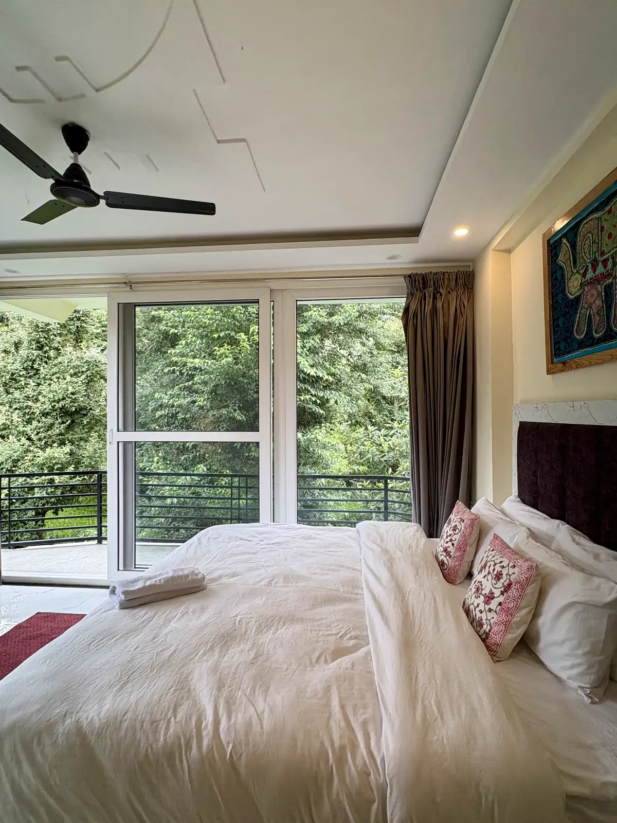 A white-linen bed with pink cushions beside full-height glass doors onto a railed balcony